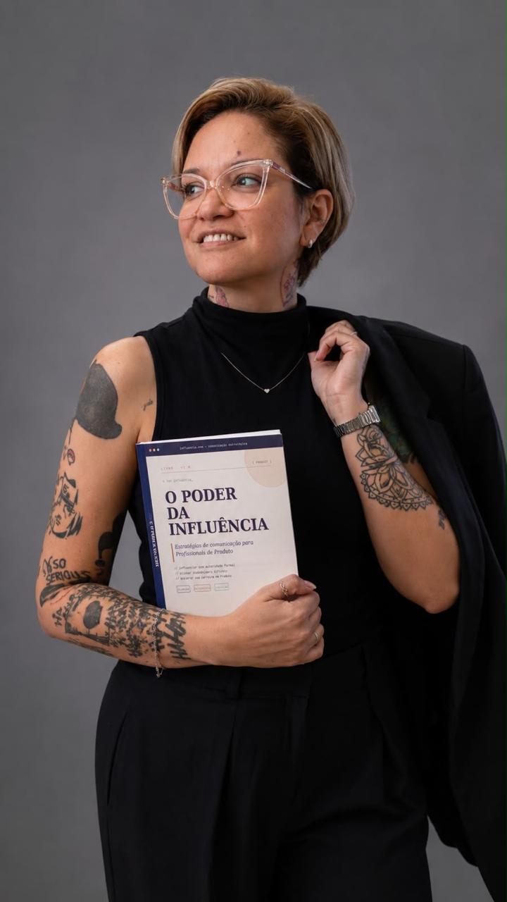 Fernanda Carvalho de blazer sobre o ombro, segurando um livro, sorrindo e olhando para o lado